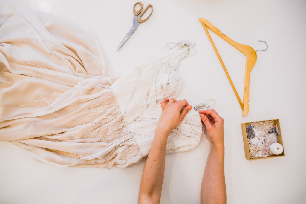Das Bild zeigt ein Tüllkleid mit weißem Top und rosafarbenem Rock. An dem Kleid hantieren zwei Hände, daneben liegen ein Kleiderbügel und eine Schere.